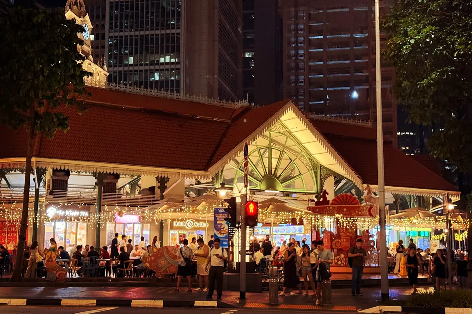Exterior night view of the historic Lau Pa Sat hawker centre in Singapore, featuring its iconic Victorian architecture illuminated under warm fairy lights, set against a backdrop of modern skyscrapers.
