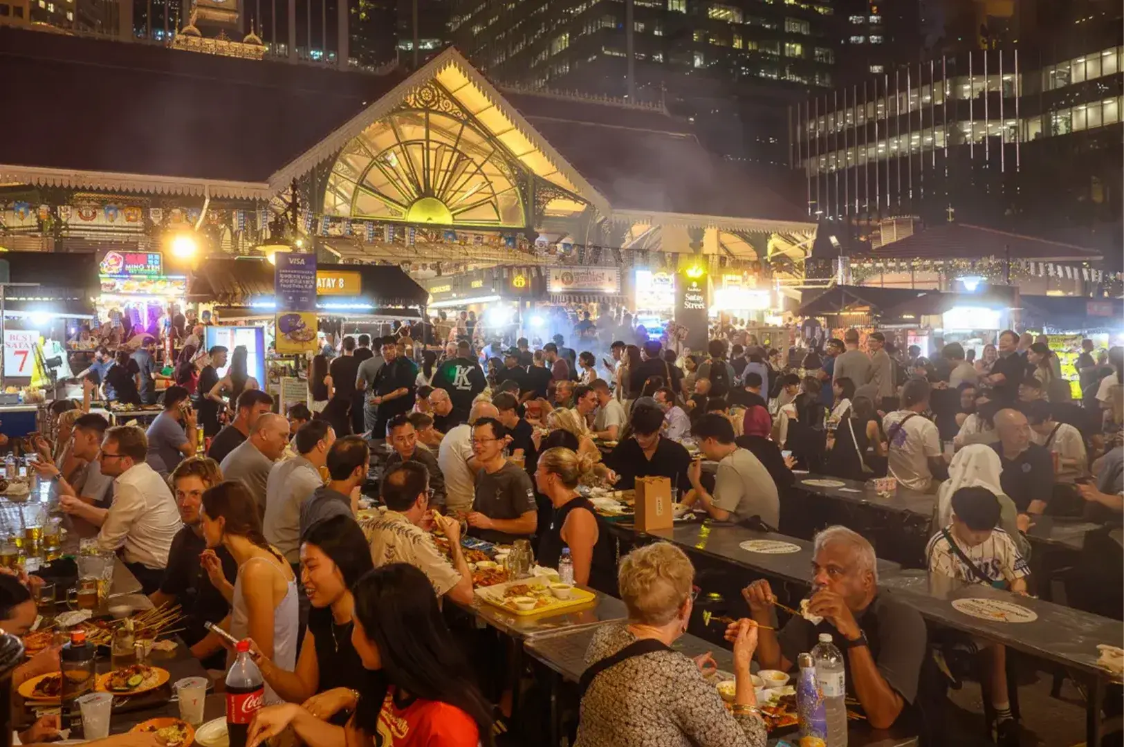 Bustling atmosphere of Satay Street at Lau Pa Sat, Singapore, filled with locals and tourists enjoying good food and a lively outdoor dinner experience surrounded by historic architecture and city skyline views.