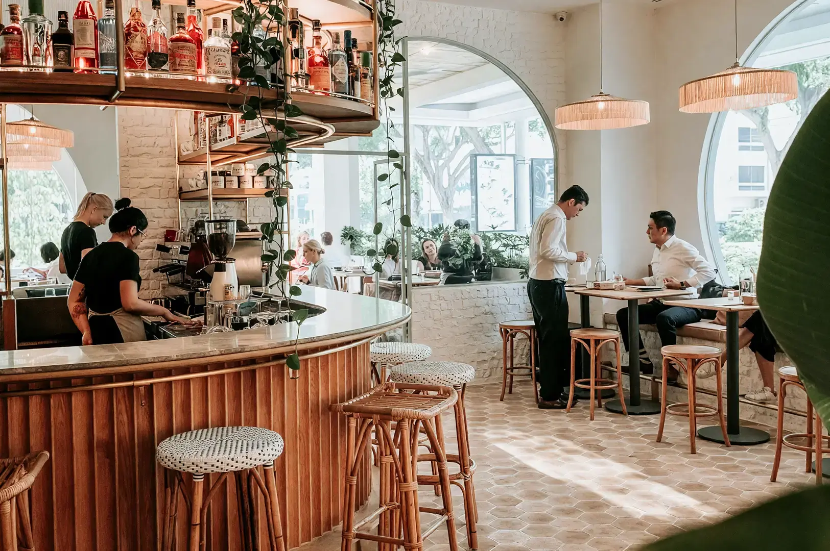 Vibrant bar area at Merci Marcel, Palais Renaissance, showcasing a rustic wooden bar, hanging greenery, and attentive staff serving guests in a sophisticated, casual atmosphere.