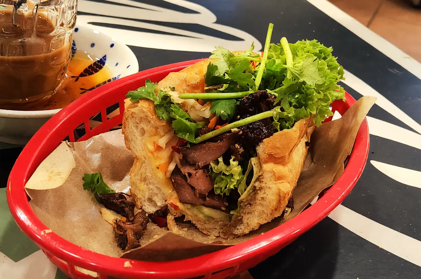 A fresh Banh Mi sandwich filled with grilled meat and cilantro, served in a red basket with a coffee, showcasing the diverse international and local food influences in Geylang.