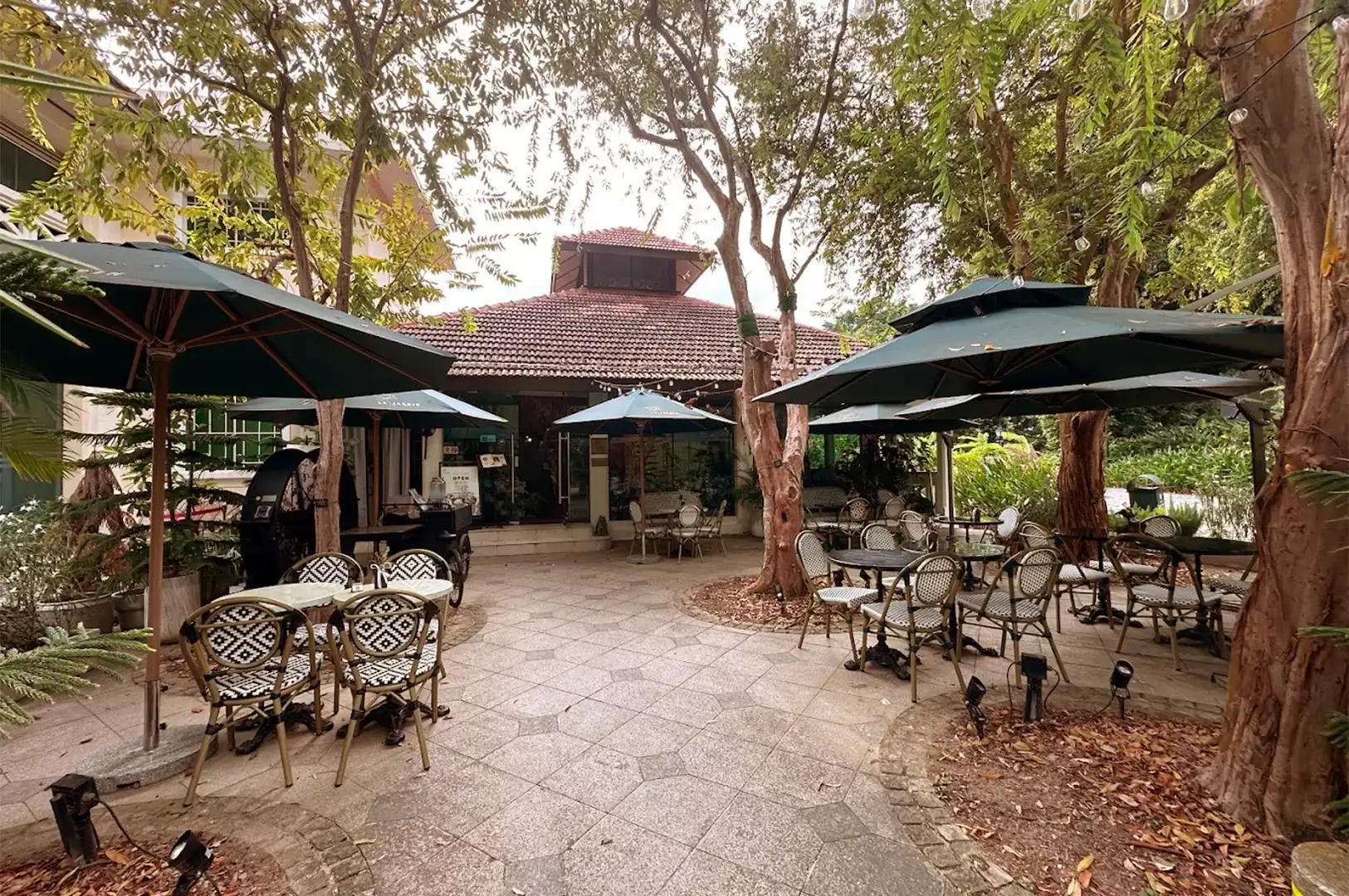 Outdoor patio seating at Le Jardin restaurant, featuring garden-style tables and chairs under trees at the Fort Canning Arts Centre, Singapore.