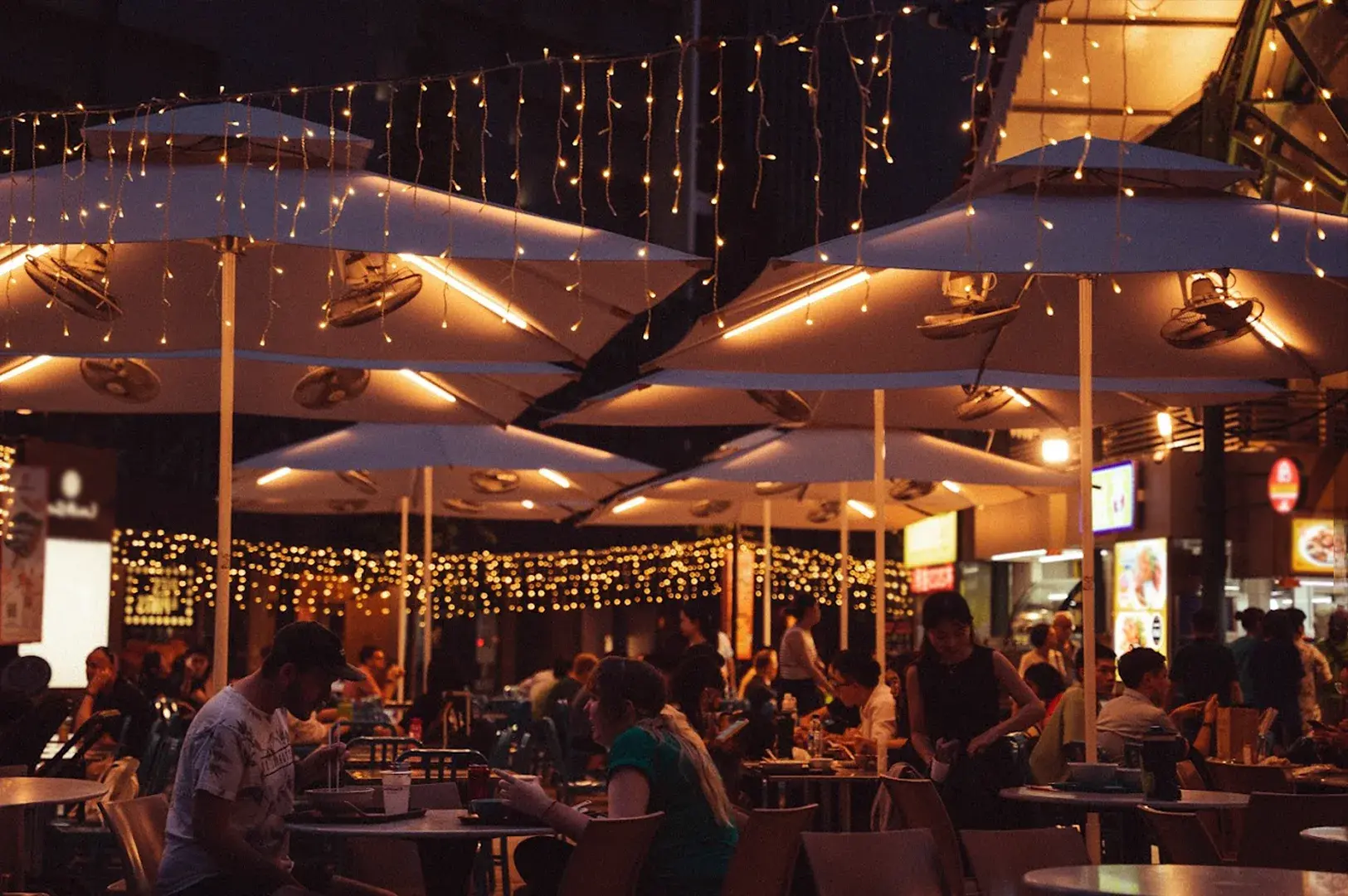 Casual outdoor al fresco dining atmosphere at Lau Pa Sat, Singapore, with patrons enjoying food under large umbrellas adorned with string lights, creating a cozy and vibrant evening dining ambiance.