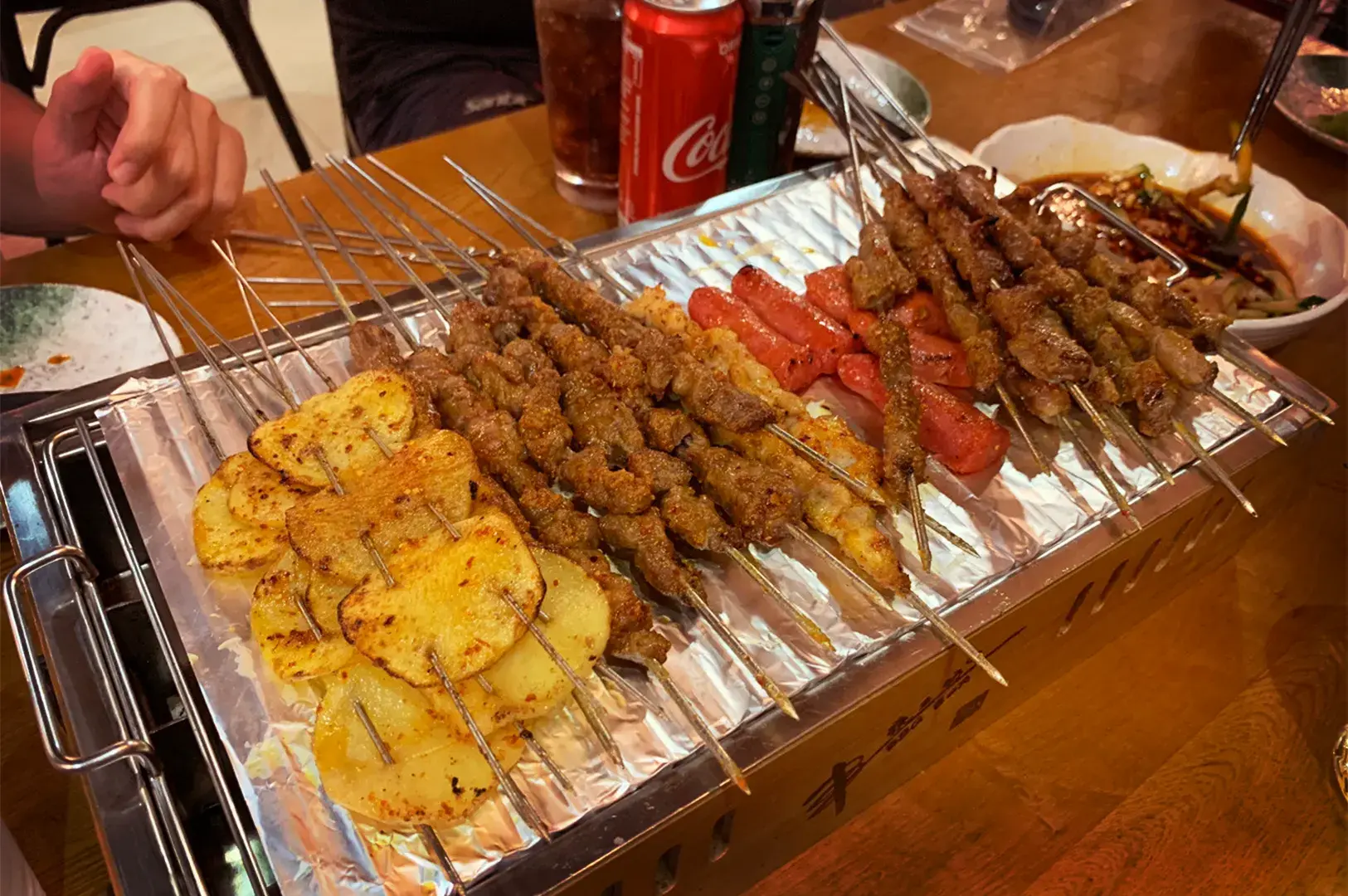 A close-up of savory grilled meat skewers sizzling on a metal tray from BBQ Box, a street food favorite synonymous with late-night hunger and supper hunts in Geylang.