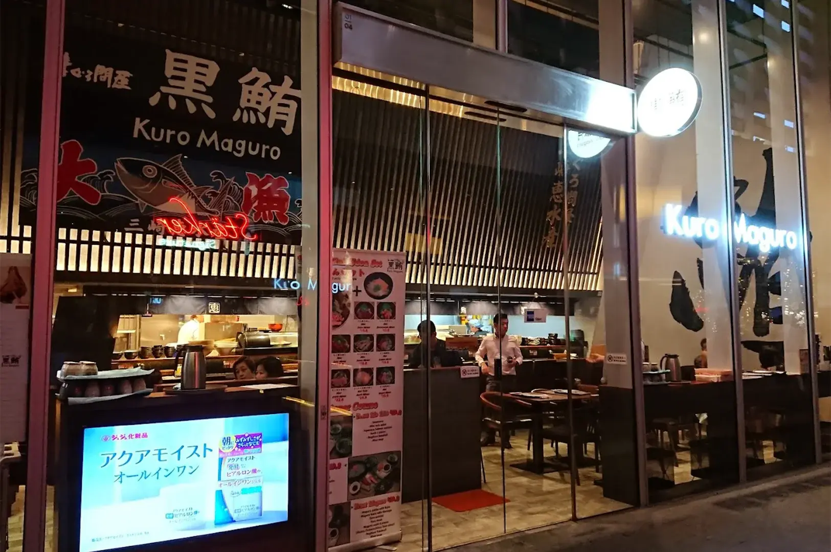 Exterior facade of Kuro Maguro Sushi and Dining at night, featuring a modern glass storefront and signage highlighting fresh tuna specialties.
