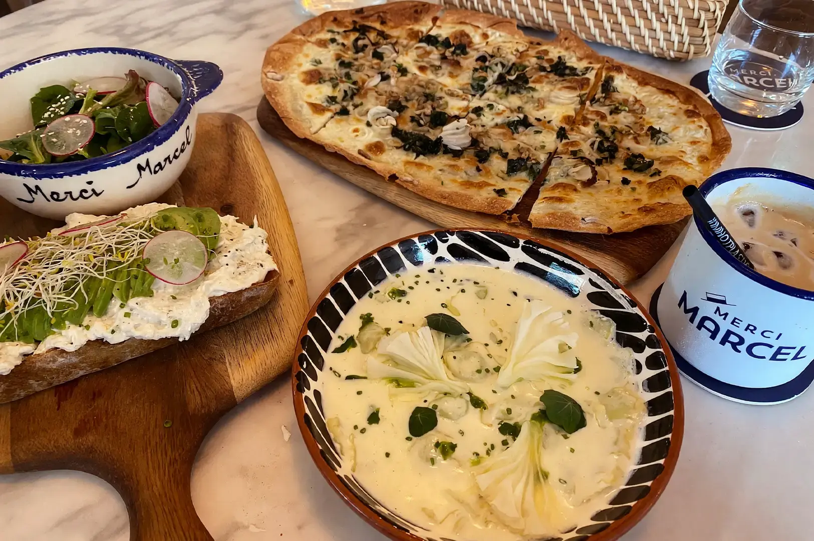 A marble table spread at Merci Marcel Orchard, featuring a plate of creamy signature Ravioles de Royans in the foreground, alongside a wild mushroom tarte flambée, avocado toast with sprouts, a side salad, and an iced latte.