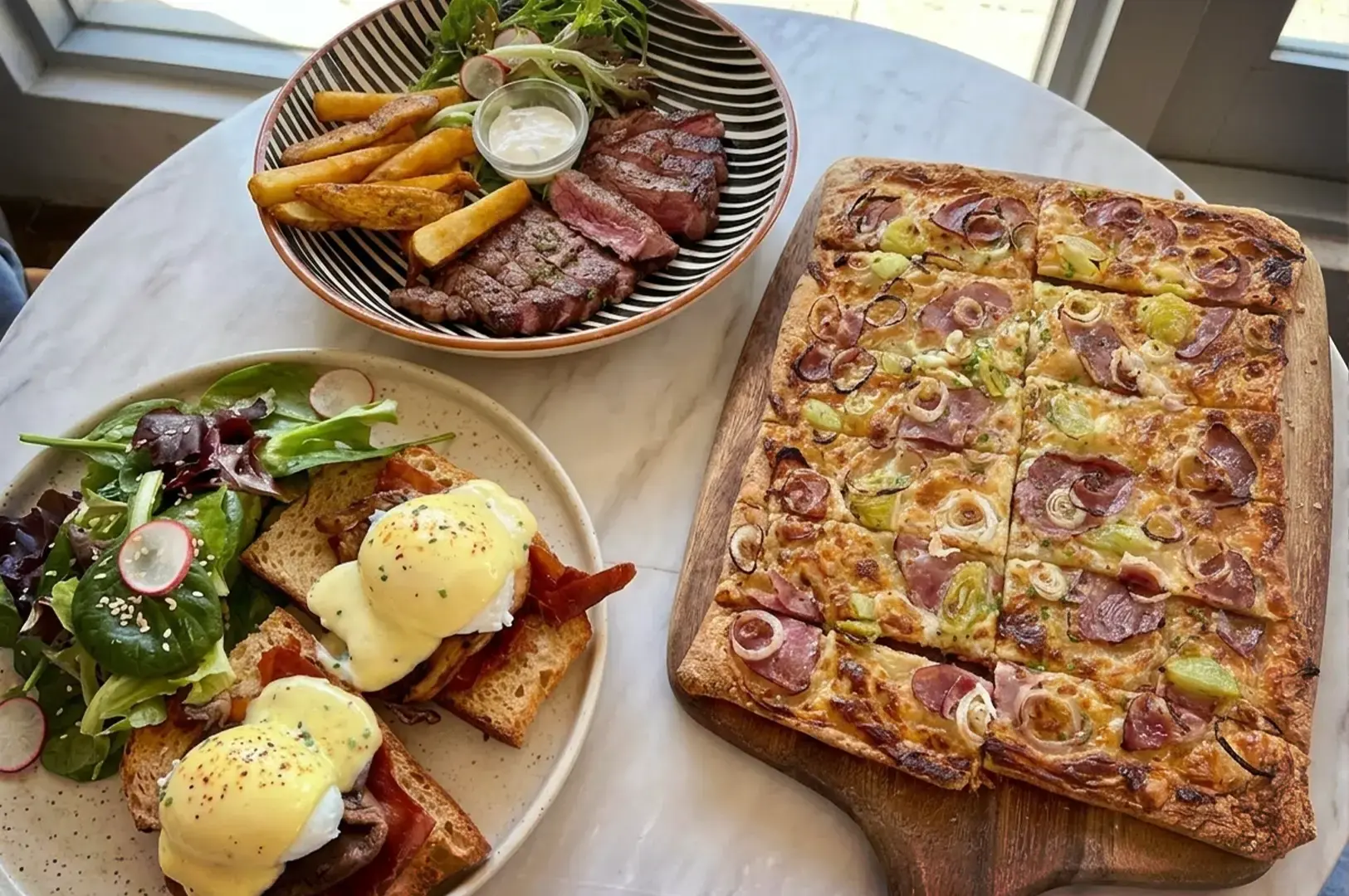 A delicious spread of French brunch favorites at Merci Marcel orchard, featuring classic Eggs Benedict with poached eggs and bacon on toast, a savory Tarte Flambée, and tender steak frites served with a side salad and dipping sauce.