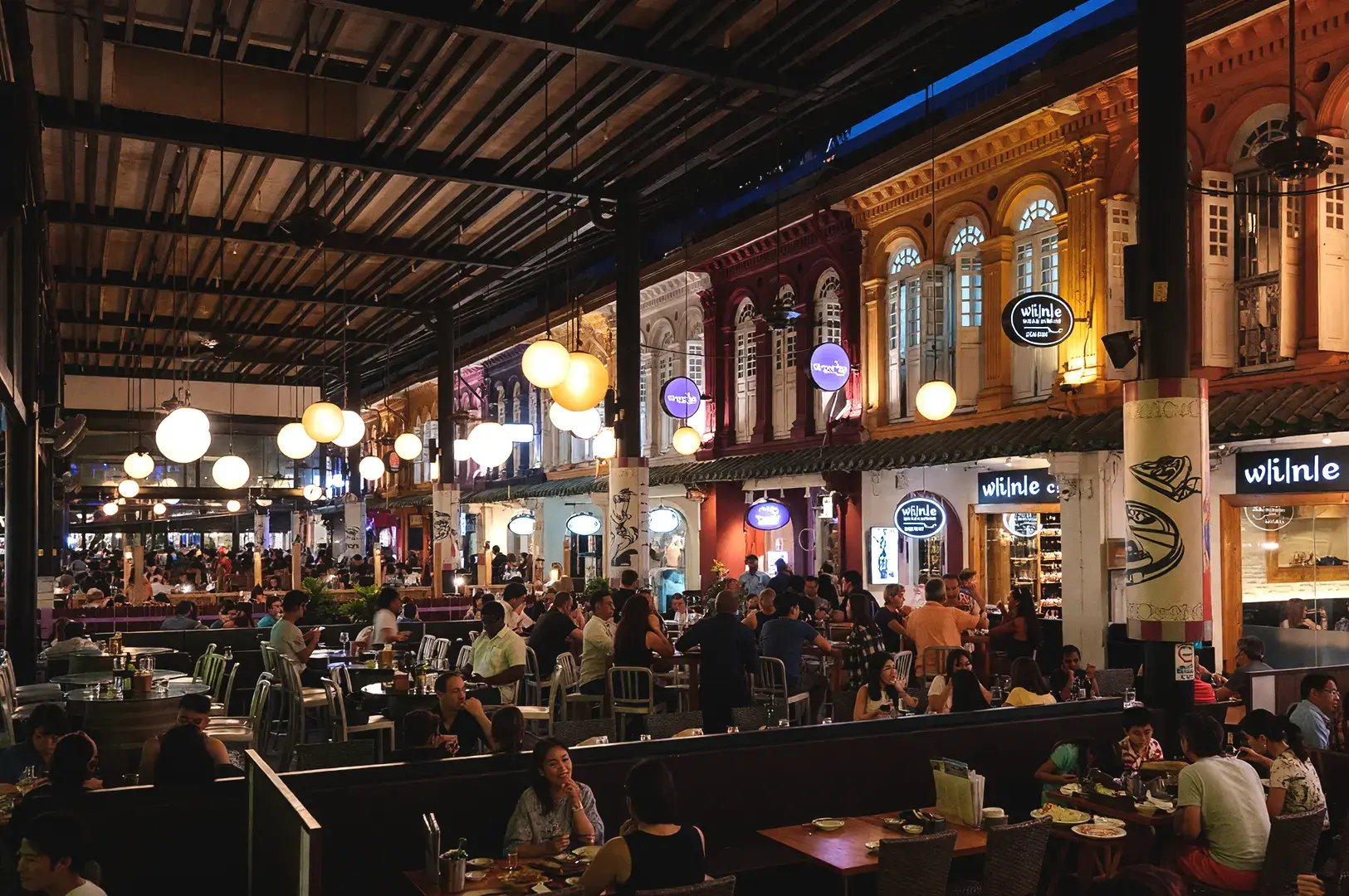A bustling night scene at a vibrant Somerset food spot, with diverse groups of friends enjoying late-night feasts at outdoor tables under hanging ball lights and illuminated traditional shophouse facades.