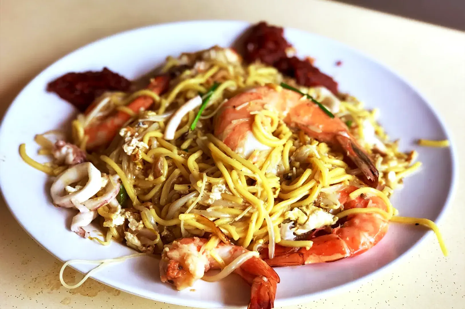 A plate of Hokkien Mee featuring fresh prawns and squid, garnished with sambal chili, representing the iconic stir-fried noodle dish available in Geylang.