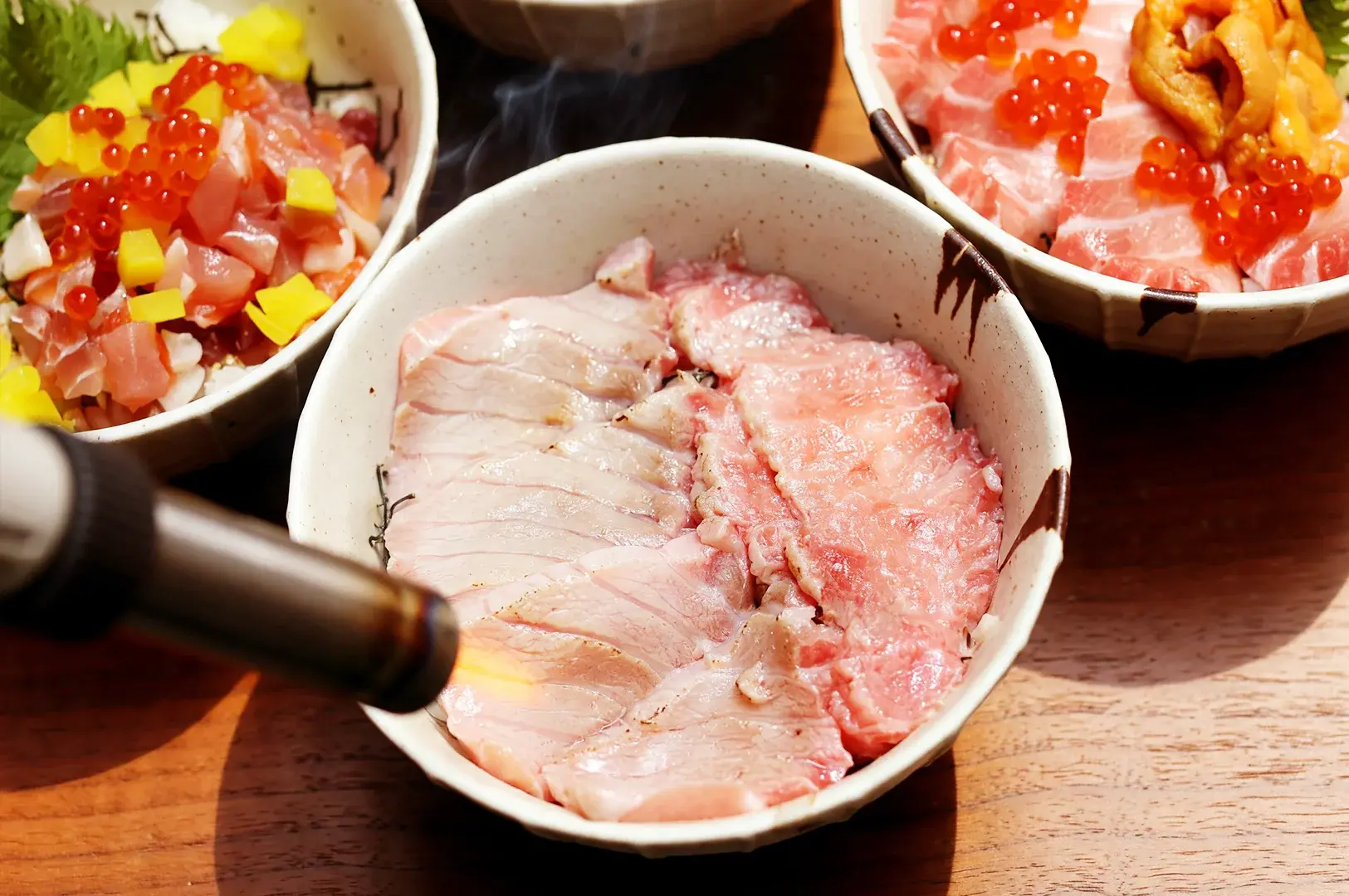 A chef using a kitchen torch to sear fresh tuna slices of the Toro Aburi Meshi Donburi bowl, served alongside the signature Maguro and Salmon Oyako Meshi Donburi bowls.