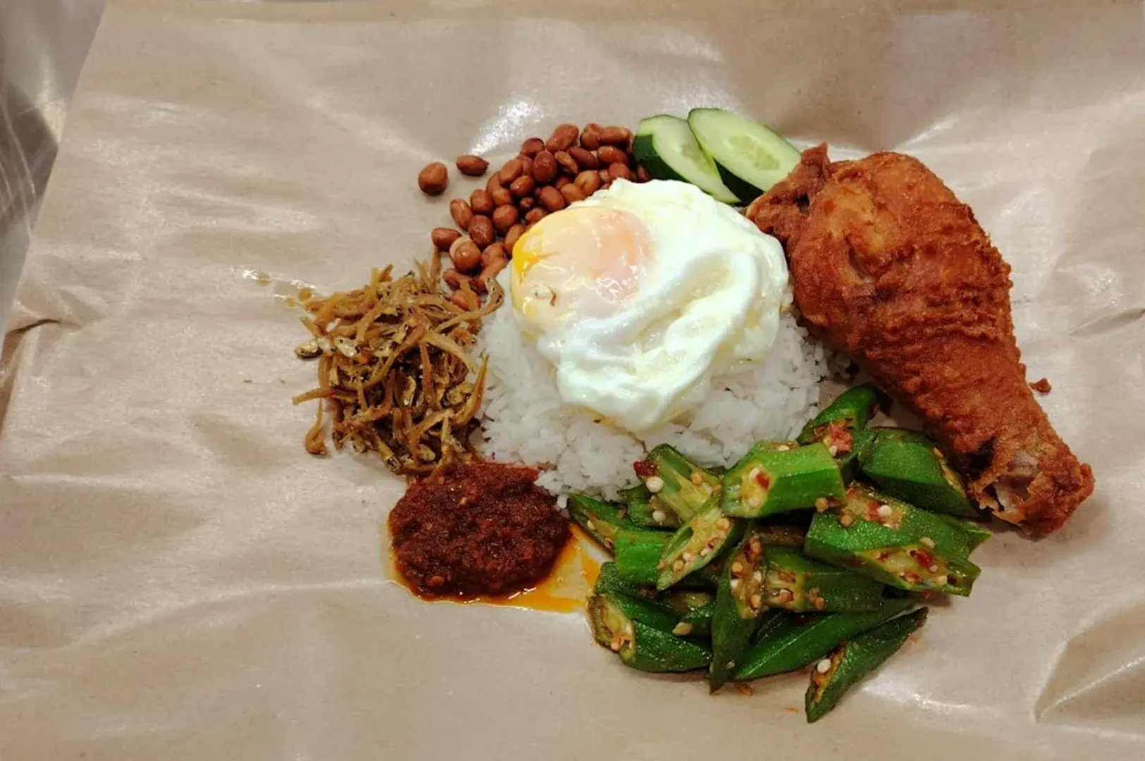 A classic Nasi Lemak set served on traditional brown paper, featuring crispy fried chicken, fried egg, peanuts, anchovies, okra, and spicy sambal—a beloved local meal in Geylang.