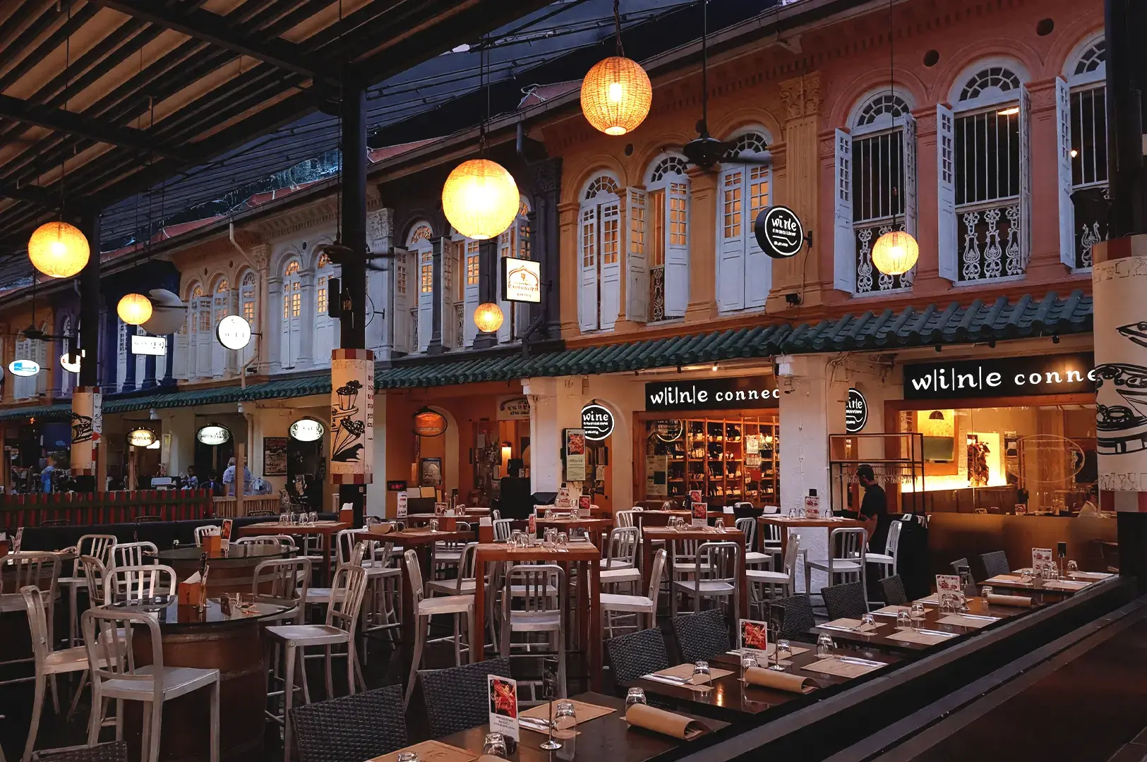 A vibrant evening view of an outdoor dining area at Somerset for late-night feasts, featuring rows of dining tables set with plates and silverware, surrounded by colorful, illuminated shophouse facades. The space is warmly lit by hanging lanterns and ambient lights, inviting friends for food.