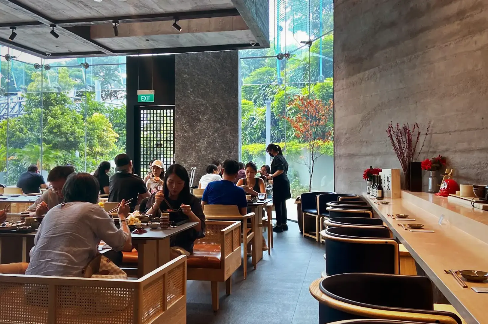 Patrons enjoying a premium lunch omakase experience inside the modern, glass-walled dining area at Ganko Sushi, featuring minimalist wooden furniture and lush garden views.