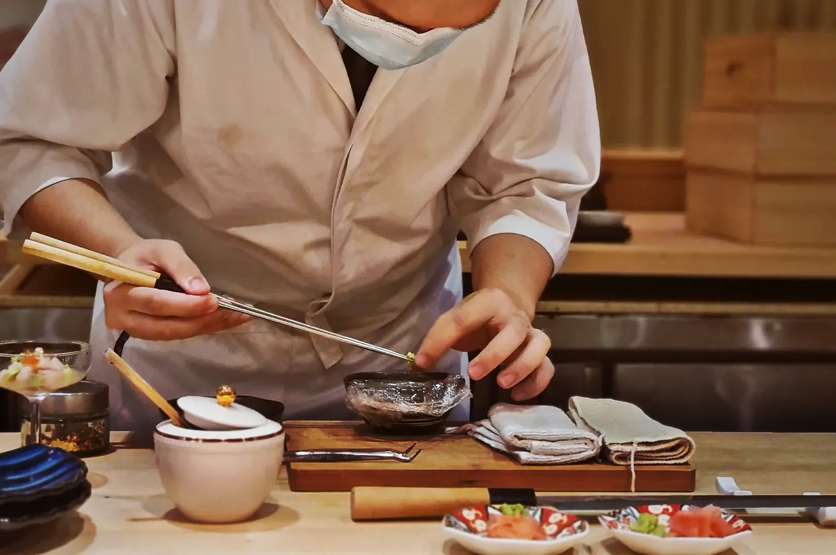A sushi chef in a mask preparing delicate ingredients with precision and balance for high-end lunch omakase sets.
