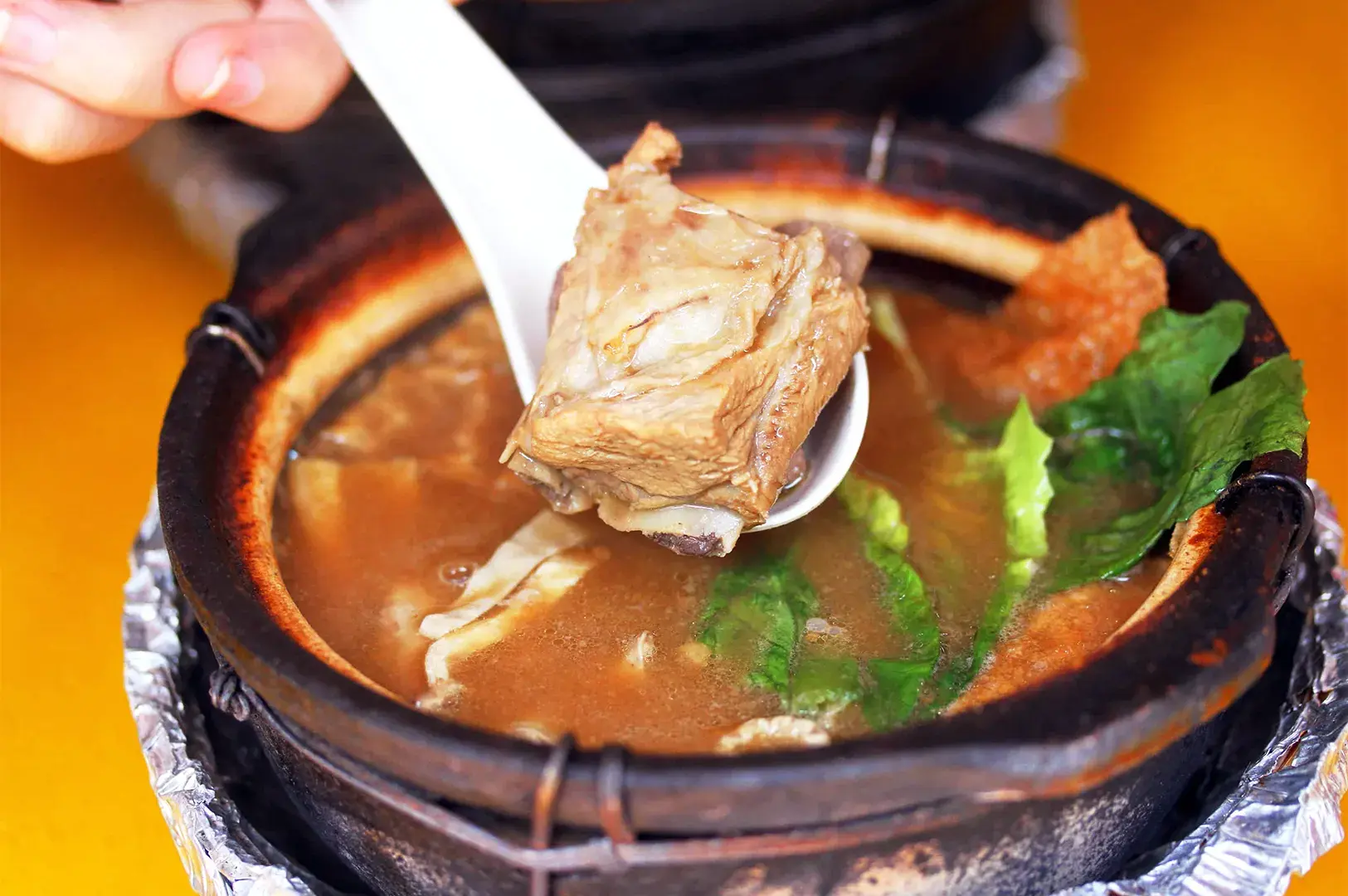A spoon lifting a tender, flavorful pork rib from a piping hot claypot of herbal Bak Kut Teh, a must-try traditional supper staple found in Geylang.