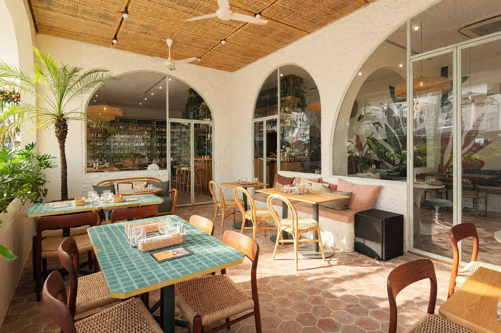 Stylish outdoor dining terrace at Merci Marcel in Palais Renaissance, featuring green tiled tables, natural rattan furniture, and arched glass windows in a chic French-inspired setting.