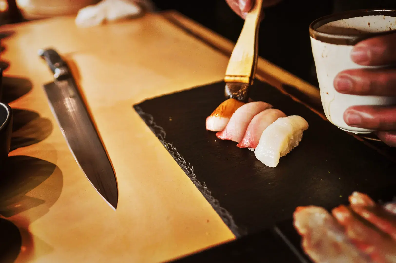 Close-up of a chef using a brush to apply sauce to fresh nigiri sushi served on a black stone plate to complete the dish during lunch omakase.