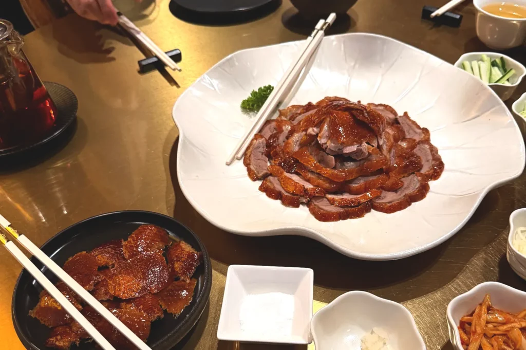 A beautifully plated serving of signature Peking duck with crispy, golden-brown skin and succulent meat at Min Jiang, Dempsey Hill, accompanied by traditional condiments and dipping sauces on a classic gold-toned restaurant table.