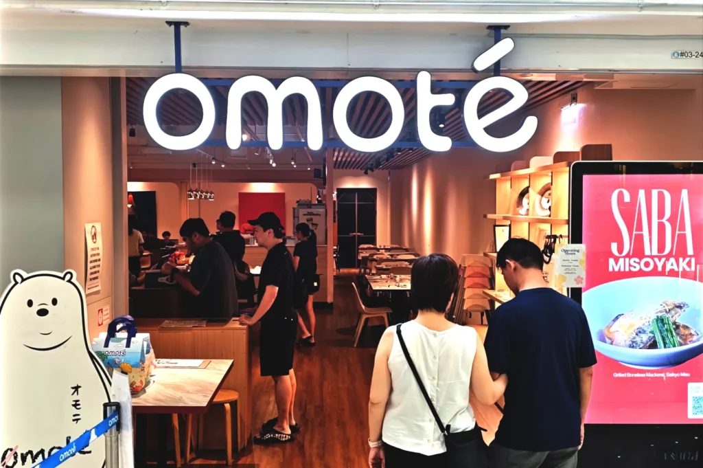 Entrance to Omote, a popular Japanese restaurant in Singapore, featuring its signature glowing signage, customer dining area, and a digital menu board highlighting fresh chirashi bowls.