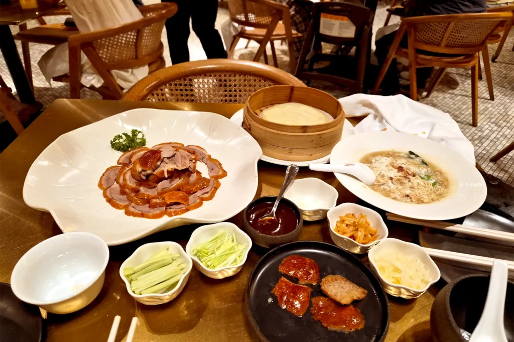 A spread of shared Chinese dishes, including Peking duck, pancakes, and noodles, set on a table at Min Jiang in Dempsey, capturing a quiet lunch focused on conversation and a sense of belonging.