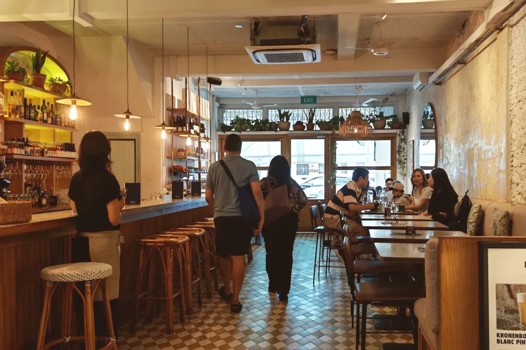 A cozy, warmly lit restaurant interior viewed from the perspective of a solo diner, showcasing a bustling bar counter, wooden tables with patrons enjoying their meals, and rustic decor details that create an inviting and memorable dining atmosphere.