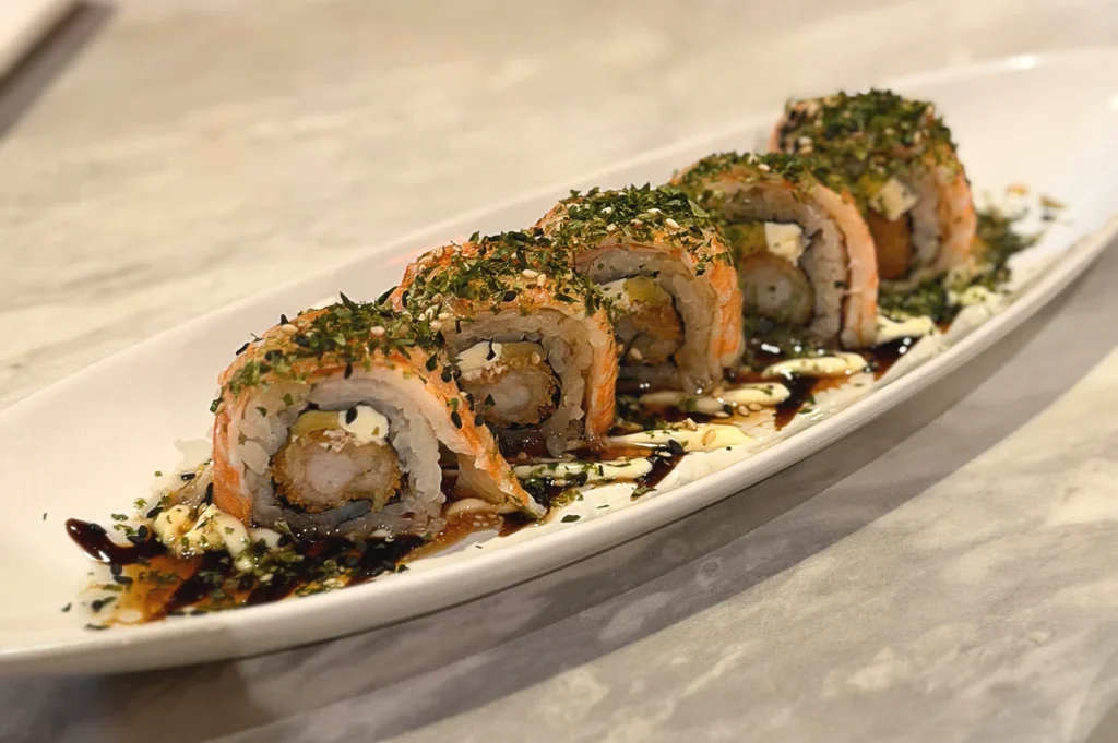 Artisan salmon sushi roll garnished with green seaweed sprinkles, beautifully plated on an elegant white dish at Omote, Singapore.