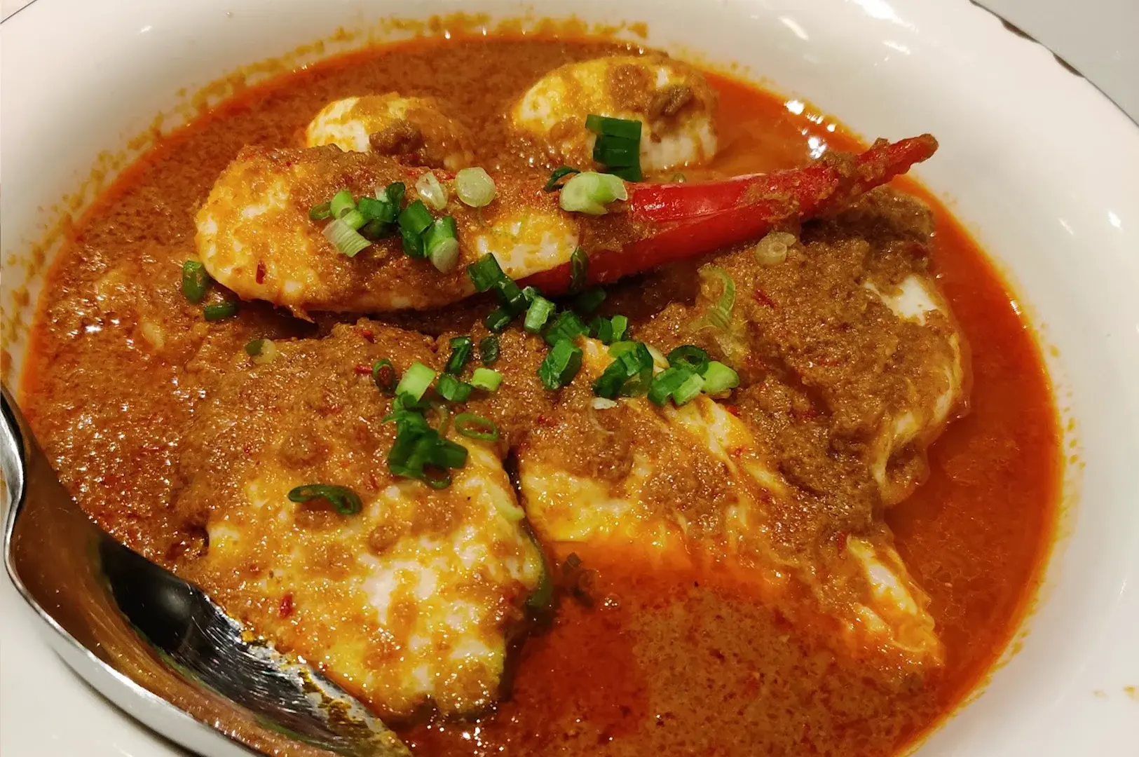 Close-up of rich, sambal prawns served at Rempapa, garnished with fresh spring onions and red chili, reflecting the depth of flavors in Chef Damian D'Silva’s heritage cooking.