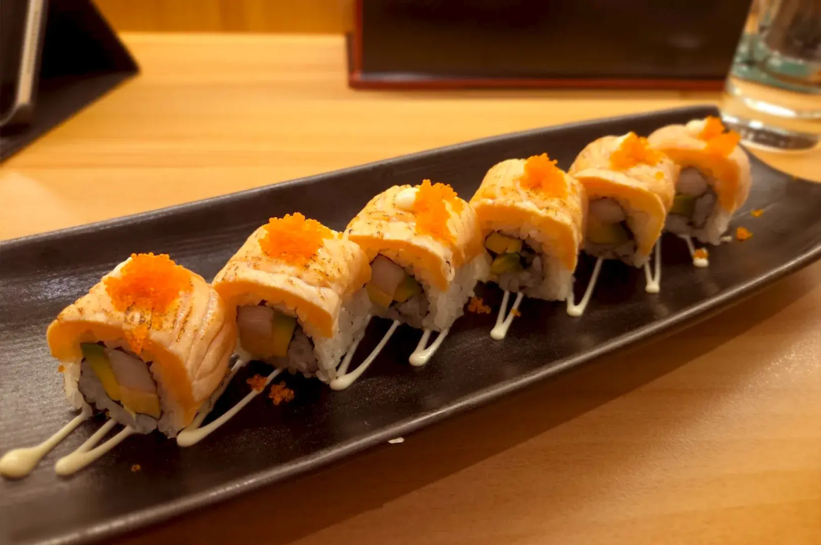 A platter of seared salmon aburi sushi rolls topped with fish roe, served on a black ceramic plate in a modern Singaporean sushi restaurant.