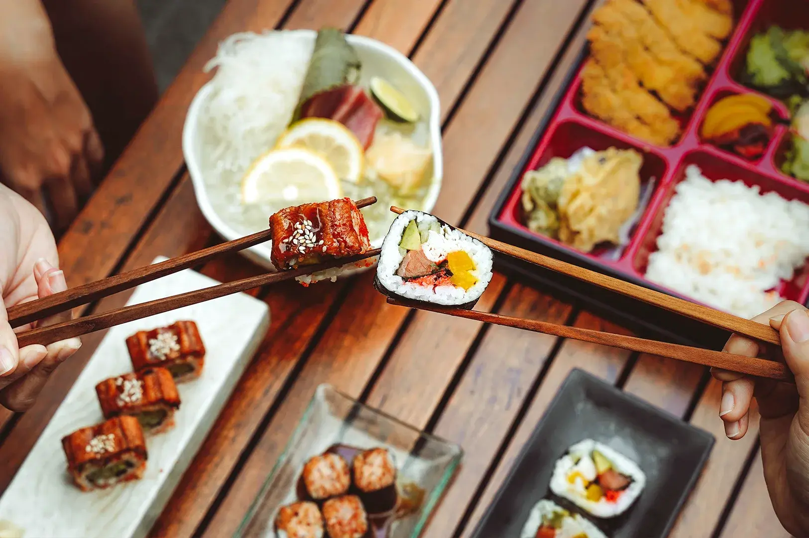 A close-up view of hands using wooden chopsticks to pick up different types of sushi rolls during a casual Japanese lunch experience in Singapore.