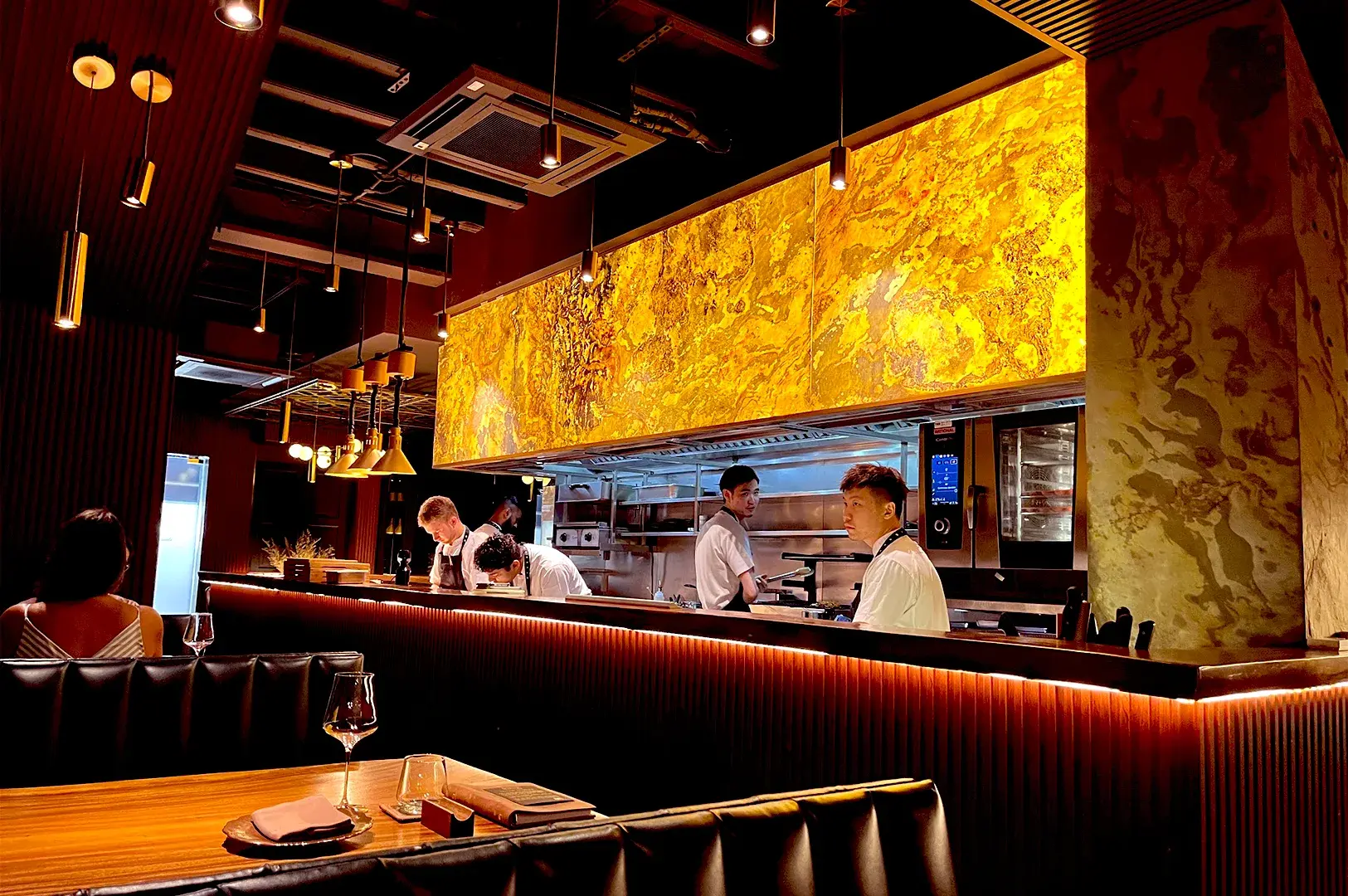 The intimate, moody dining atmosphere at Thevar Singapore, featuring a sleek open kitchen where chefs prepare modern Indian tasting menus under warm, ambient lighting.