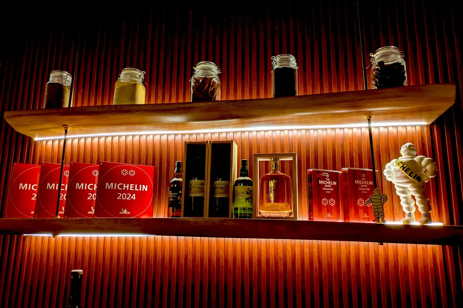 A shelf at Thevar Singapore showcasing prestigious Michelin Guide awards from 2023 and 2024, alongside premium spirits and ingredients, highlighting the restaurant's culinary excellence.