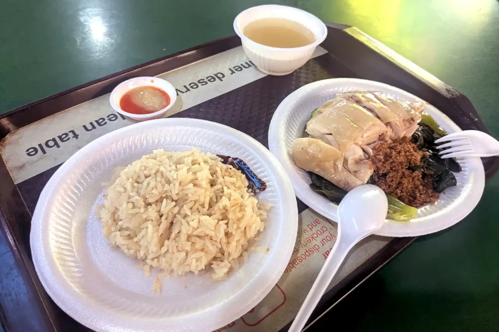 A traditional Singaporean hawker meal on a tray featuring chicken rice, a bowl of soup, and chili sauce, highlighting the value of affordable local dining.
