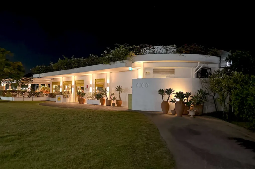 Exterior night view of Fico Singapore, a seaside Italian restaurant with illuminated white architecture, potted plants, and a manicured lawn under a dark sky.