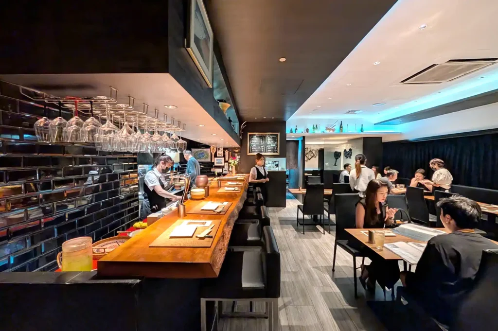 A wide interior view of Kazu Sumiyaki Singapore featuring a warm wooden counter bar, contemporary dark styling, and diners enjoying smoky yakitori nights.