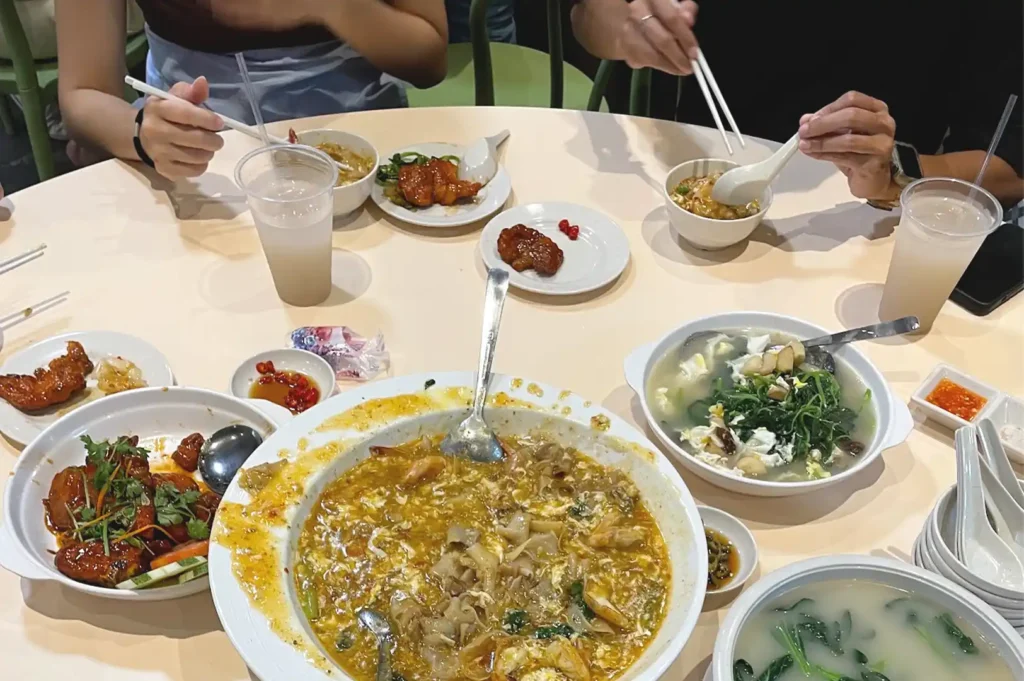 A top-down view of friends gathered around a table filled with comforting local dishes and drinks in Singapore, enjoying a rare phone-free dinner centered on meaningful conversation and shared meals.