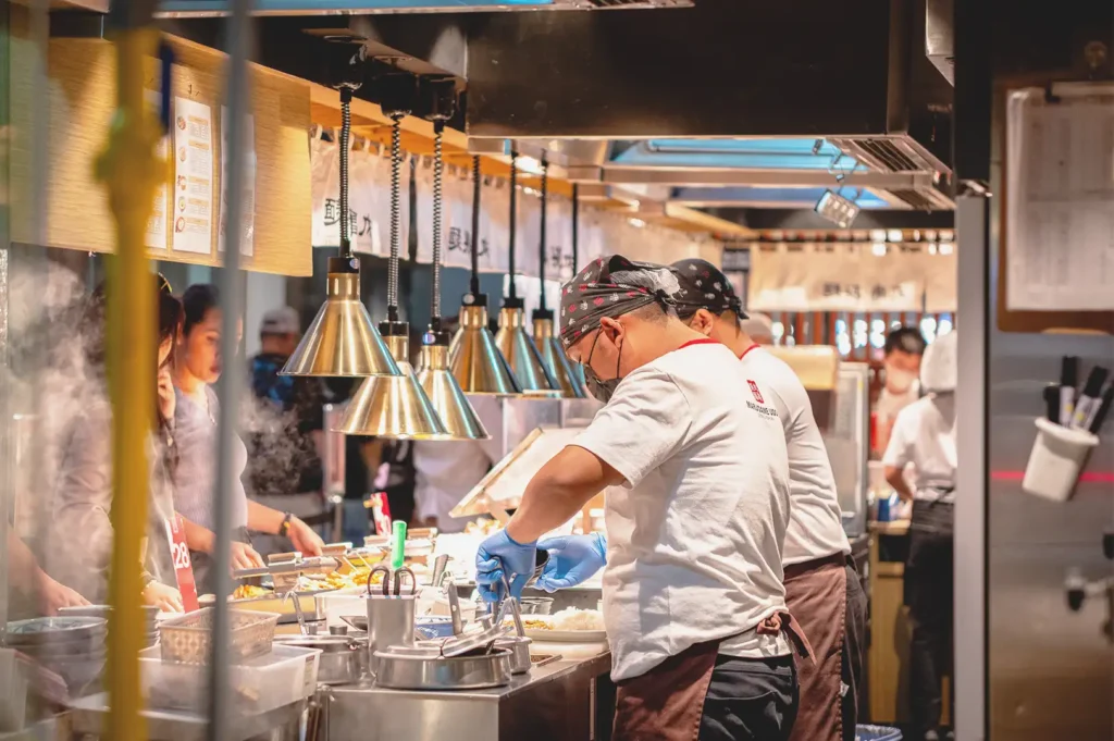 A busy restaurant kitchen in Singapore with chefs focused on food preparation under warm hanging lights, reflecting the hidden human effort, time, and stories that shape the true value of a meal.