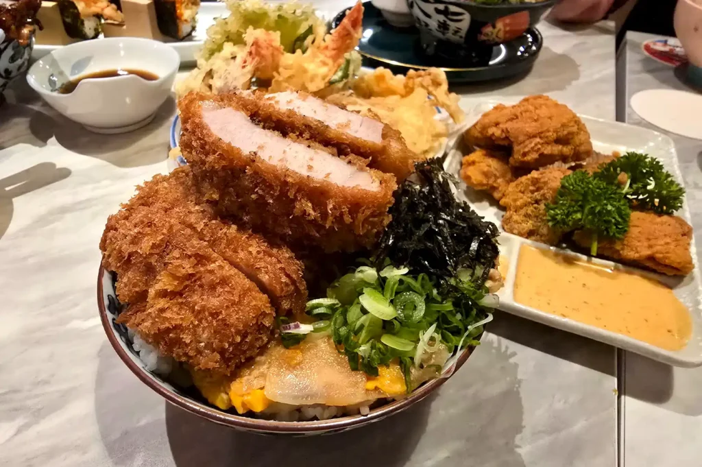 A hearty bowl of Japanese katsu don topped with thick-cut, rustic fried pork cutlets, sliced onions, egg, and green seaweed, served alongside fried chicken and dipping sauce, celebrating the authentic and memorable experiences of a polished meal.