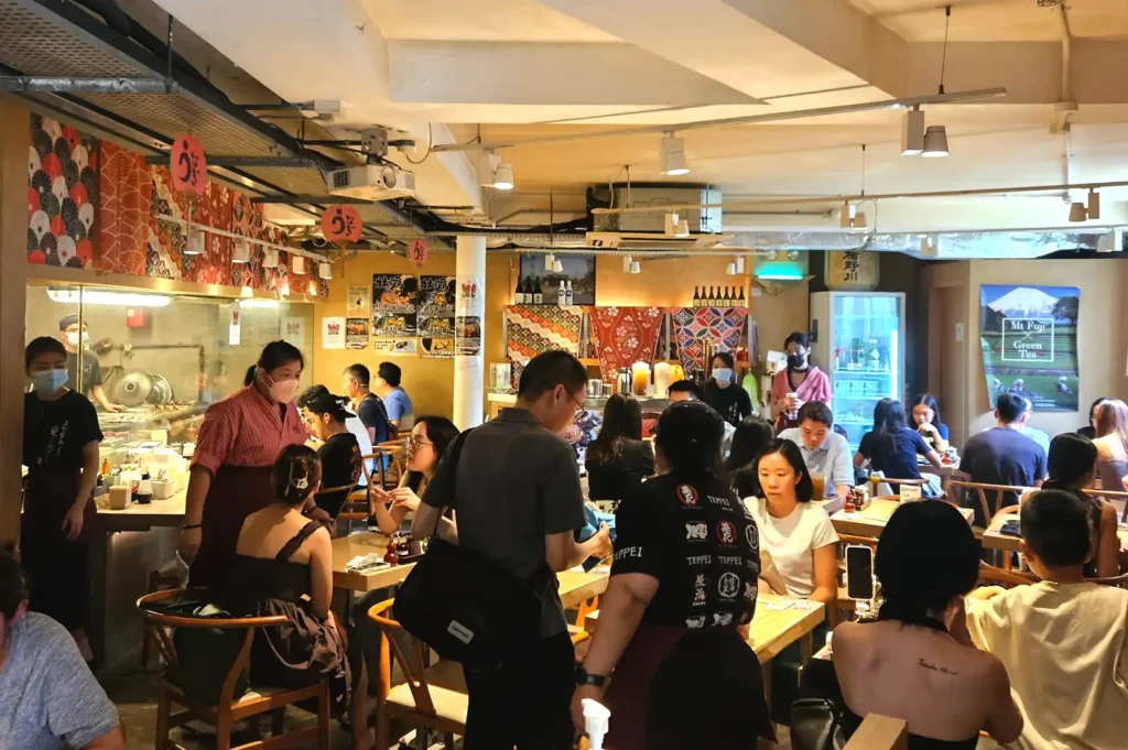 A bustling restaurant scene in Singapore with patrons waiting in line and seated at tables, capturing the patience, shared friendships, and stories behind the local dining culture.