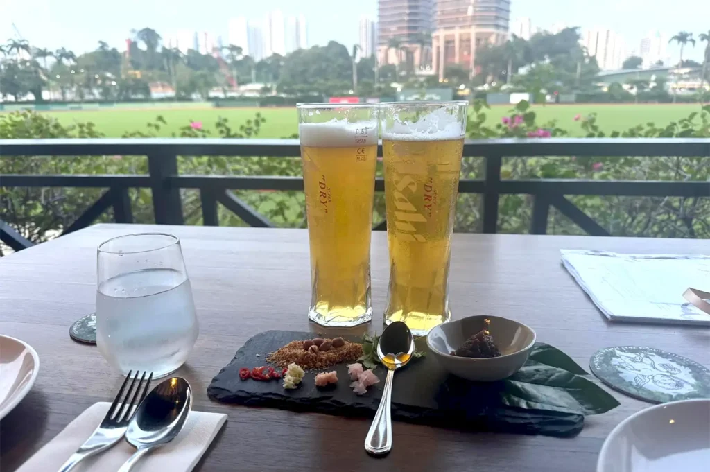 Two tall glasses of Asahi beer and a slate board of snacks on an outdoor restaurant table overlooking a lush green field, capturing an unranked dining experience meant to be remembered.