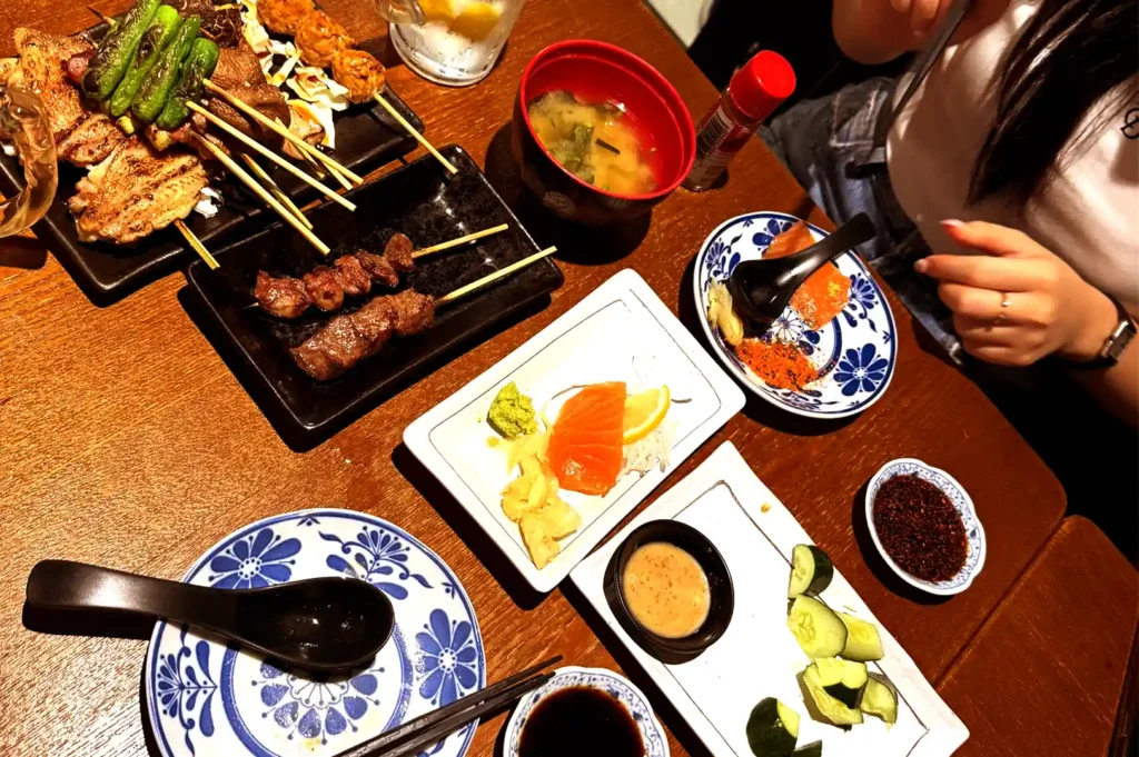A top-down view of a wooden dining table laden with Japanese food items including yakitori skewers, salmon sashimi, sliced cucumber, and miso soup, inspiring conversations that continue long past the meal.
