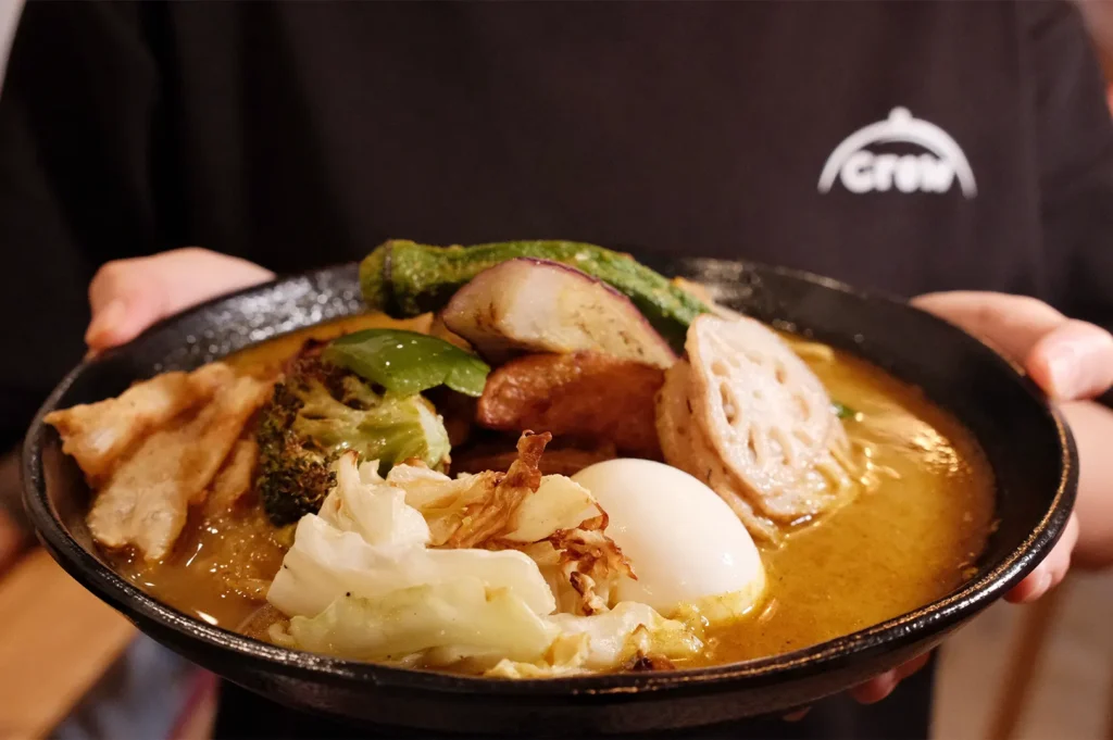 A person holding a bowl of Soup Curry by Ki-setsu at Fortune Centre, showcasing aromatic Hokkaido soup curry laden with assorted vegetables and a soft-boiled egg.