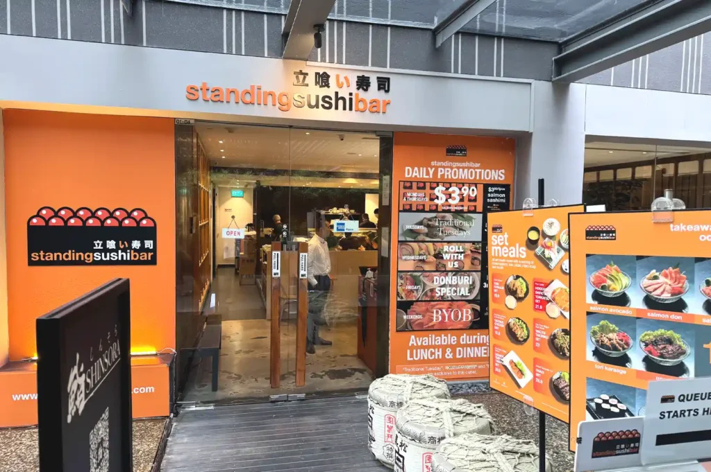 Exterior storefront of Standing Sushi Bar showcasing bright orange signage, promotional banners, daily set meal displays, and traditional barrels.
