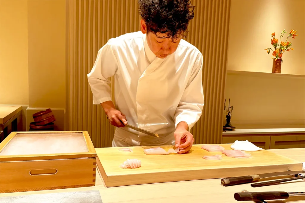 Chef Sakuta in a crisp white uniform meticulously slicing raw fish on a wooden cutting board at Sushi Sakuta, a two-Michelin-star omakase restaurant in Singapore.