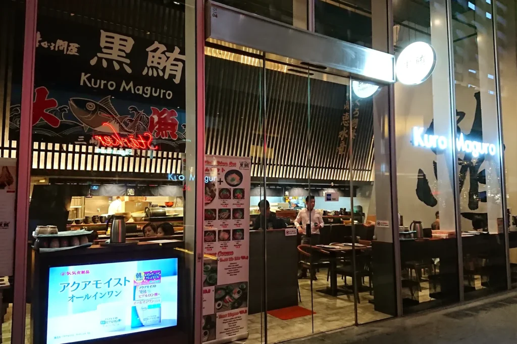 The modern glass-fronted exterior of Kuro Maguro restaurant in Singapore, showcasing Japanese signage, an illuminated menu stand, and a view into the dining area.