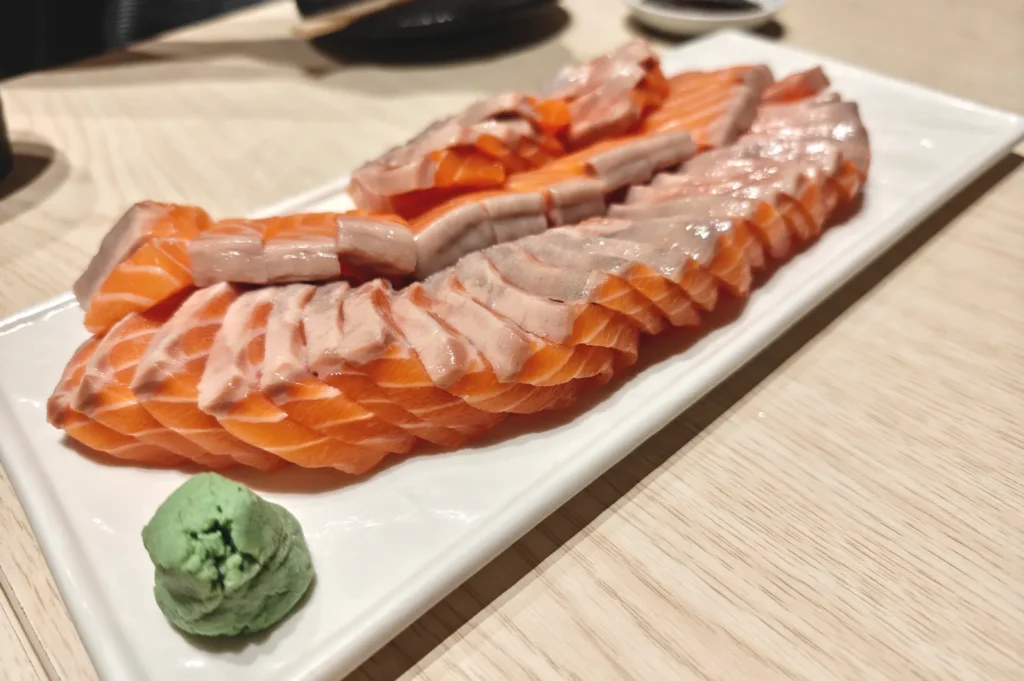 A white rectangular plate featuring neatly sliced fresh salmon sashimi arranged in rows alongside a mound of green wasabi.