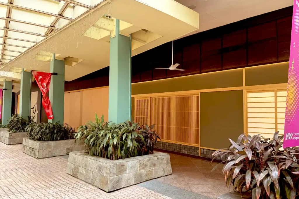 The exterior facade of Sushi Sakuta featuring traditional Japanese wood panels, neutral tones, and lush green potted plants along the walkway.