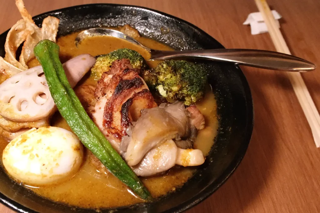 A bowl of Hokkaido soup curry from Soup Curry by Ki-setsu highlighting tender chicken pieces, broccoli, okra, and a whole egg resting in a light, spiced broth.