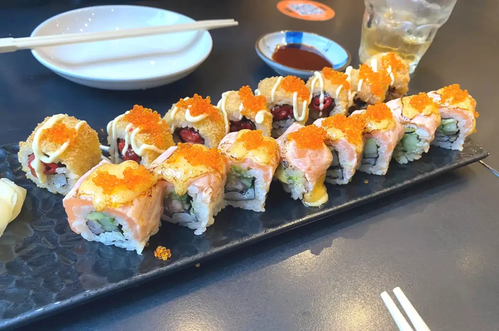 A black rectangular plate serving an assortment of gourmet maki sushi rolls topped with melted cheese, spicy sauce, and orange fish roe.
