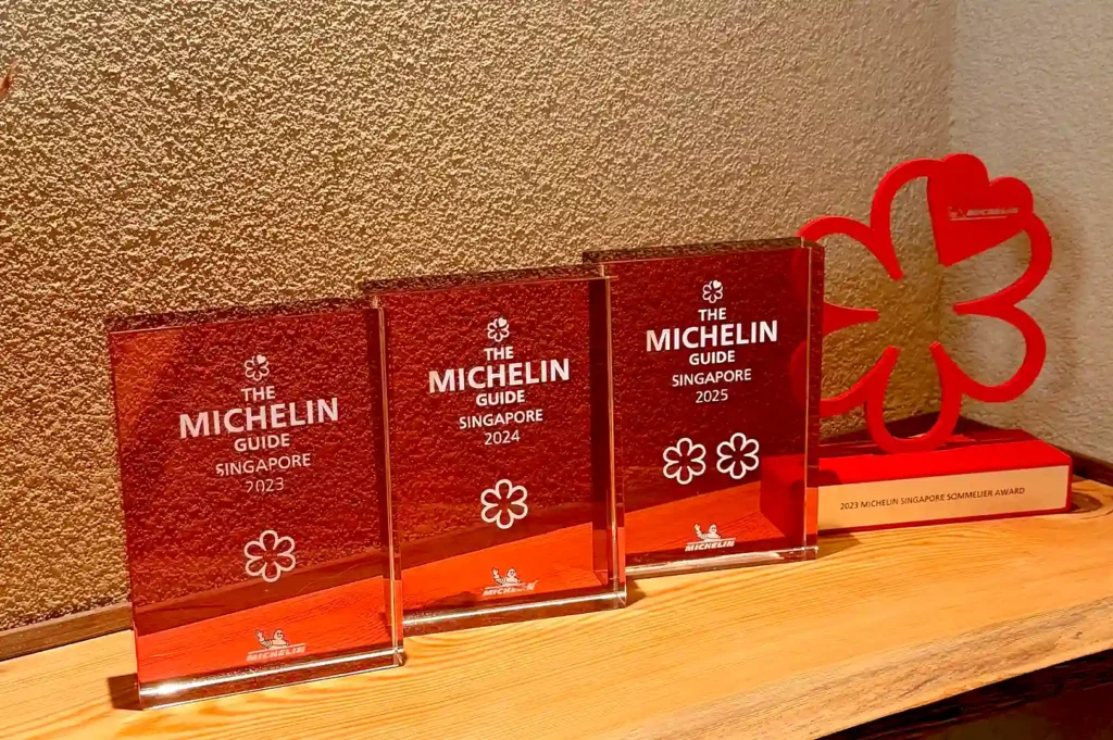 A collection of official Michelin Guide plaques and awards displayed on a wooden shelf, highlighting Sushi Sakuta’s prestigious two-Michelin-star achievements in Singapore.