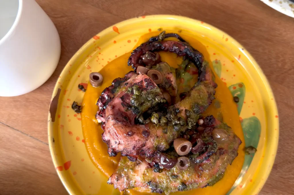 Tender grilled octopus tentacles served over a vibrant orange puree, garnished with olives and capers on a yellow ceramic plate at Fico Singapore.