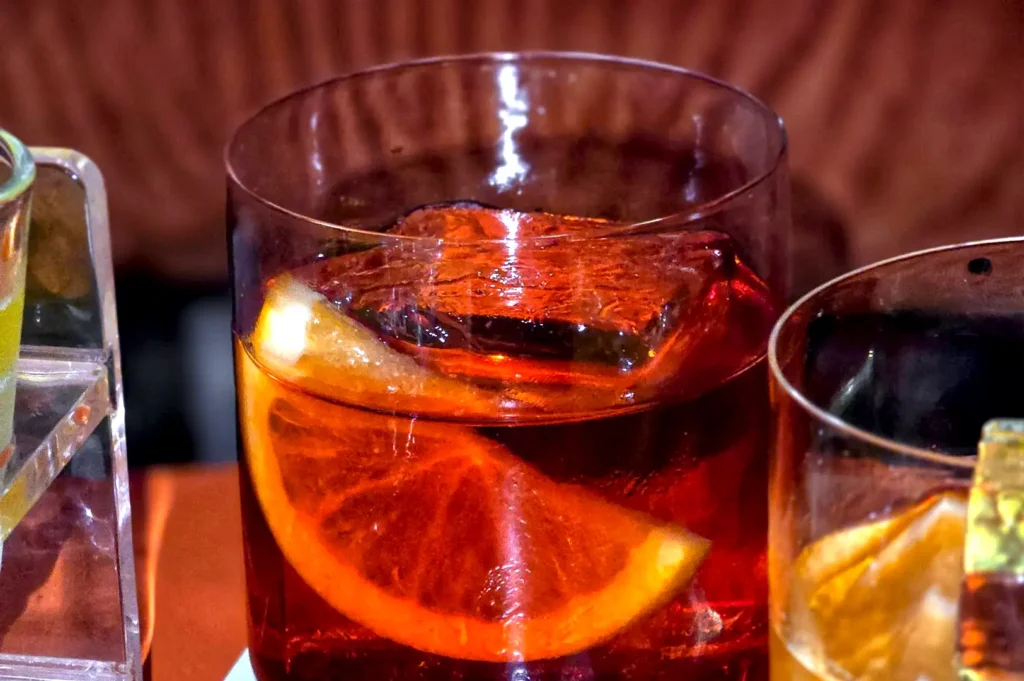 A close-up shot of a signature Lapsang Souchong Negroni in a short glass with a large clear ice cube and an orange slice garnish at Rasa Space.